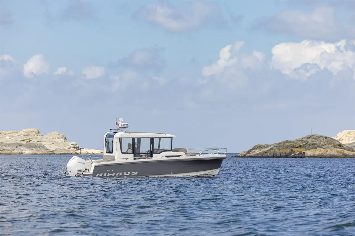 A Nimbus boat navigates calm waters near rocky shores under a blue sky with clouds, showcasing its sleek design.
